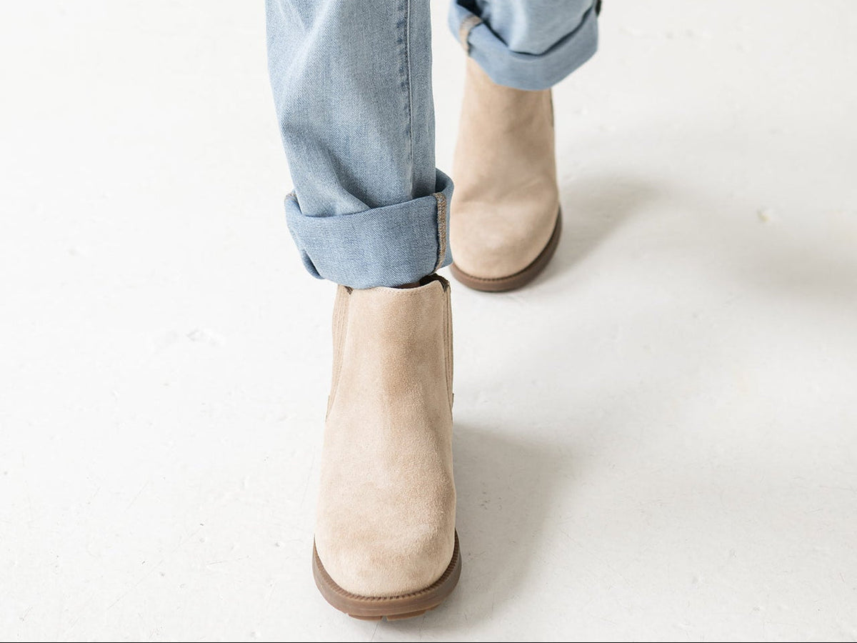 Beige suede ankle boots worn with light blue cuffed jeans, shown from the knees down on a clean white studio floor.
